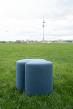 Two blue cylindrical ottomans on grass with a distant cityscape.