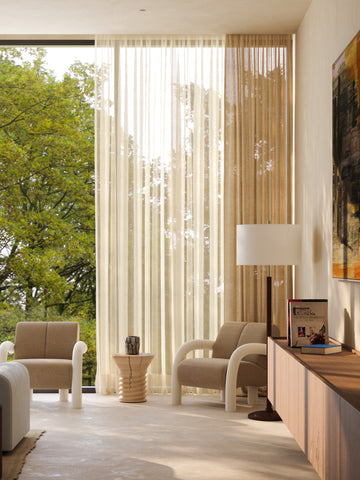 Modern living room with beige chairs, a side table, and a large window with sheer curtains.